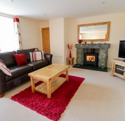 A living room with a brown leather sofa a wooden coffee table on a red rug a stone fireplace and a TV on a wooden stand at Tyn Y Buarth in Dyffryn Ardudwy
