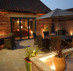 An outdoor patio with wicker chairs tables an umbrella large potted plants and a brick house with glass doors at Sykes Lodge in Malton