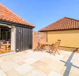 An outdoor patio with a wooden table and four chairs next to a black wooden building with open doors showing a living room at Sparrow Barn in Reepham