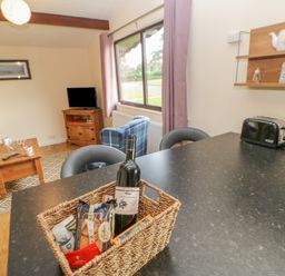 A kitchen counter with a basket containing a wine bottle and snacks in a living room with a wooden coffee table blue armchair and a TV on a wooden stand at Orchid Lodge in Saltburn-by-the-Sea