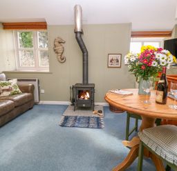 A living room with a brown leather sofa a wood burning stove a round wooden table with flowers and wine glasses at Higher Lampra in Mullion