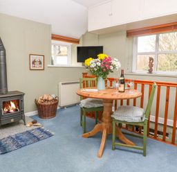 A dining area with a round wooden table with flowers and wine glasses near a wood stove with fire at Higher Lampra in Mullion