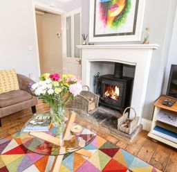 A living room with a glass coffee table, colorful rug, armchair with yellow patterned pillow, fireplace with wood stove, and a TV on a wooden stand at The Cottage at Broadway in Broadway