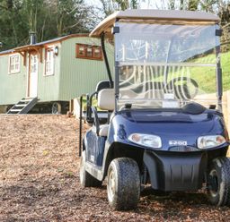 A golf cart parked on a wood chip path next to a green shepherd hut with windows and stairs in a wooded area at Little Silver Leaf in High Bickington