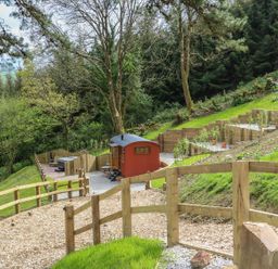 A wooden fenced pathway leading to a small red cabin on a hillside with trees and greenery at Little Silver Owl in High Bickington