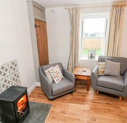 A living room with two gray armchairs a wooden side table a window with striped curtains and a wood burning stove at Coeden Bach in Burry Port