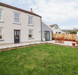 A backyard with green grass wooden patio furniture potted plants a white barbecue and a two-story house at Coeden Bach in Burry Port