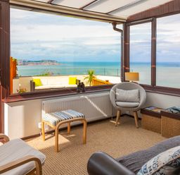 A sunroom with chairs and a footstool overlooking a balcony with a sea view at Sea-Prize View in Old Colwyn