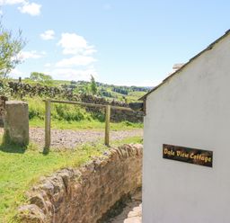 Exterior side of a white building with a Dale View Cottage sign and a stone wall with green fields in the background at Dale View Cottage in Allendale near Hexham