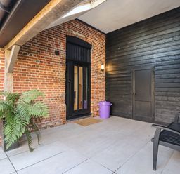 A covered outdoor patio area with brick and black wooden walls a black door a black chair a potted plant and a purple trash bin at Grange Barn in Gedding near Felsham