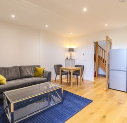 A living room with a grey sofa with yellow cushions a glass coffee table a blue rug a small dining table with two chairs a lamp on a cabinet a refrigerator and wooden stairs at Grange Barn in Gedding near Felsham