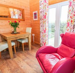 A dining area with a wooden table white chairs a red leather armchair and floral curtains by glass doors at Borah Lodge in Mousehole