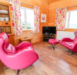A wooden living room with two red chairs a matching footstool a wooden cabinet floral curtains and a television at Borah Lodge in Mousehole