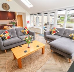 A living room with two gray sofas and pillows a wooden coffee table with flowers and large windows at Meadowside in Mousehole