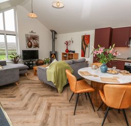 An open plan living area with grey sofas a wood burning stove a round dining table with orange chairs and a kitchen with red cabinets at Meadowside in Mousehole