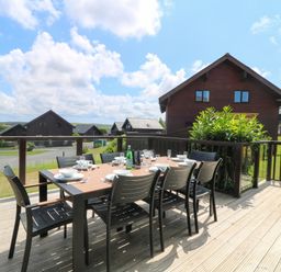 An outdoor dining table with chairs on a wooden deck overlooking houses and greenery at Willow Retallack in Retallack near St Columb Major