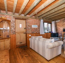 A living room with wooden beams on the ceiling a wooden door a window with curtains striped sofas a bookshelf and a TV at The Dairy Barn in Toftrees near Fakenham