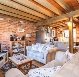 An open living room and kitchen with exposed wooden beams brick walls and light grey sofas at The Dairy Barn in Toftrees near Fakenham