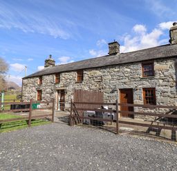 A stone cottage with three chimneys and wooden doors behind a fenced yard at Cwm Yr Afon Mawr in Llanbedr near Harlech