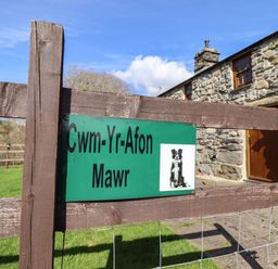 A wooden gate with a green sign showing a dog and the text Cwm Yr Afon Mawr in front of a stone building with wooden door and windows at Cwm Yr Afon Mawr Llanbedr near Harlech