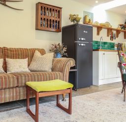 A living area with a striped sofa beside a green cushioned stool and a dark fridge near a kitchen counter at Courtyard Cottage Poplar Farm Barn Sweffling near Saxmundham