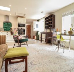 A living space with a striped sofa green cushioned stool wooden chair kitchen cabinets oven green tiled backsplash table with yellow chairs and window light at Courtyard Cottage Poplar Farm Barn Sweffling near Saxmundham