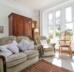 A sitting room with two upholstered armchairs a wooden cabinet a standing lamp a wooden chair and large windows at The Old Vicarage in Tiverton
