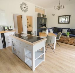 An open-plan kitchen and living area with a kitchen island, dining table with chairs, sofa, coffee table, and wall clock at The Barn in Droitwich Spa