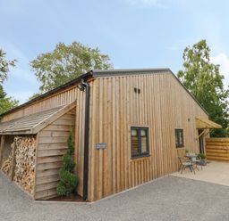 A wooden exterior building with stacked firewood on the side a small patio with two chairs and a table and trees in the background at The Barn in Droitwich Spa
