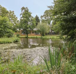 A pond surrounded by trees and plants with a wooden building in the background at The Barn in Droitwich Spa