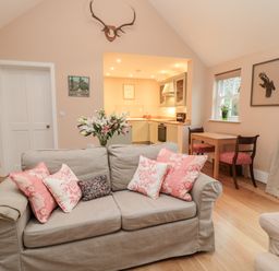 A living room with a sofa with pink cushions a small wooden table with two chairs and an open kitchen area at The Garden Cottage in West Woodburn