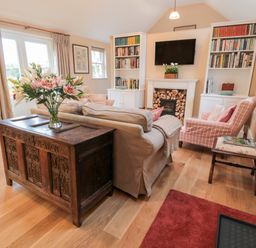 A living room with a sofa a wooden chest with flowers a fireplace with stacked firewood bookshelves and armchairs at The Garden Cottage in West Woodburn