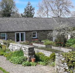 A stone cottage with a slate roof surrounded by raised garden beds and a stone wall at The Garden Cottage in West Woodburn