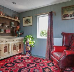 A room with a red leather armchair a wooden cabinet with shelves and copper kettles a window with a curtain and a red patterned rug on the floor at Crows Cabin in Gloucester
