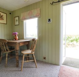 A small dining area with a wooden table and two wooden chairs near a window and an open door at Crows Cabin in Gloucester