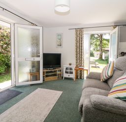 A living room with a grey sofa and striped cushions a television on a stand and open doors leading outside at Tranquillity Rosecraddoc Lodge near Liskeard