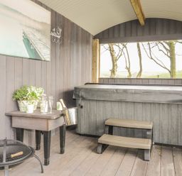 A covered outdoor area with a hot tub a small table with plants champagne glasses and a champagne bucket and a fire pit at High Rigg Shepherd's Delight in Brampton Cumbria