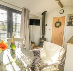 A dining area with a glass table a bottle of champagne and flowers near a window and a wood stove at High Rigg Shepherd's Delight in Brampton Cumbria