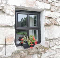 A stone wall with a black framed window and a flower pot with red flowers on the windowsill at An Teach Ban in Rossaveel County Galway