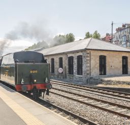 A steam train at a railway platform next to tracks and a stone building at Evening Star @ Engine Shed in Whitby
