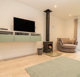 A living room with a wall-mounted television a wood-burning stove a round armchair and a glass door at Kober House in Mullion