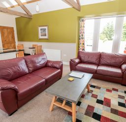 A living room with two red leather sofas a coffee table a multicolored rug and a dining table with chairs at Hemmel Cottage in Chatton