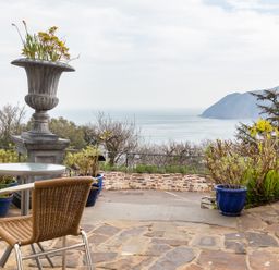 An outdoor patio with a round table and two wicker chairs overlooking the sea and hills with potted plants and a large decorative urn at Bayview Terrace in Lynton