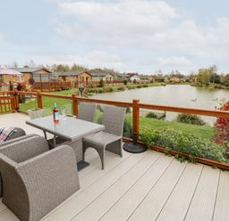 An outdoor deck with wicker furniture and a glass table overlooking a lake with houses in the background at Lake View Lodge in Marston near Barkston