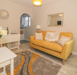 A living room with a yellow sofa a rug a small white table with flowers and an archway to a kitchen at Holly Cottage in Criccieth