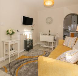 A living room with a yellow sofa a small dining table a wall-mounted tv and a doorway to a kitchen at Holly Cottage in Criccieth
