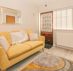 A yellow sofa with patterned pillows and a blanket in a room with a wooden door with a sailboat curtain and a window with blinds at Holly Cottage in Criccieth