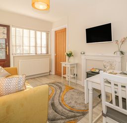 A small living room with a yellow sofa a dining table with chairs a wall-mounted tv and a window with blinds at Holly Cottage in Criccieth