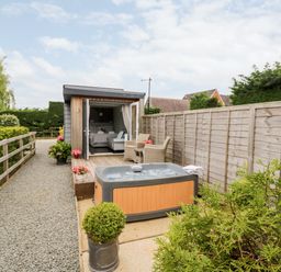 A small wooden cabin with an open door showing a bed inside a garden with a hot tub and outdoor chairs at Hydrangea Bretforton