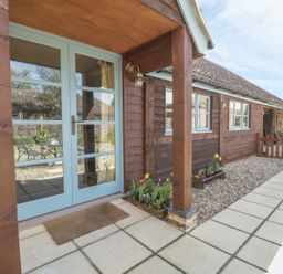 A wooden house exterior with light blue double doors and flower beds on the side at Shippon Barn in Yeovil